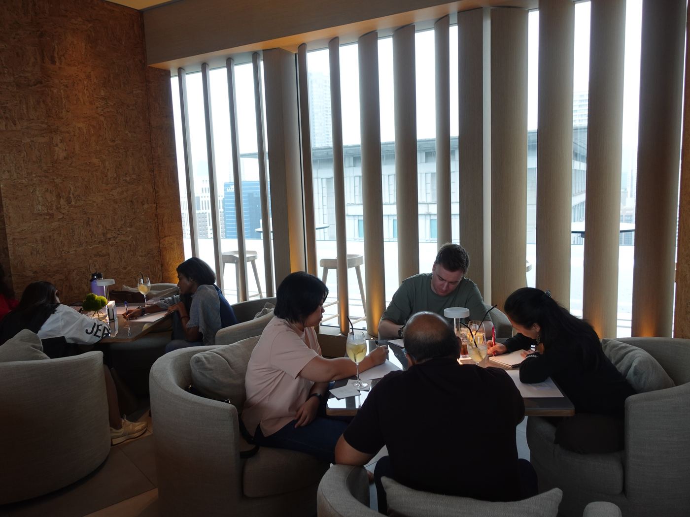 Guests sit around low tables in a lounge with tall windows, writing and talking