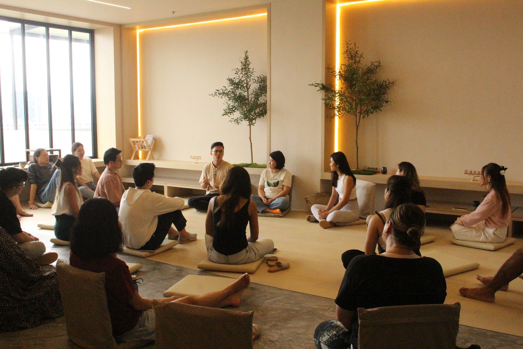 A group sits in a circle on floor cushions in a warm, softly lit room, listening to a speaker
